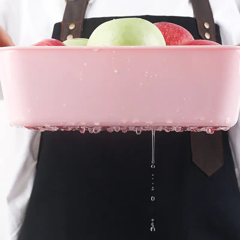 Pink Multifunctional Sink Chopping Board Drain Basket with apples being rinsed, held by a person wearing a black apron.